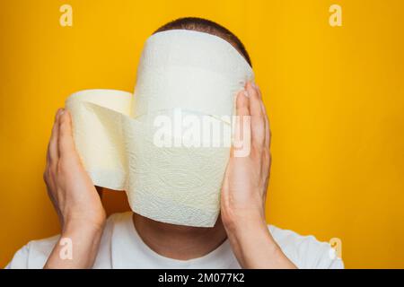 Un uomo in una camicia blu gli avvolse il volto in carta igienica, cercando di proteggersi dal coronavirus. In studio su sfondo giallo. Protezione Foto Stock