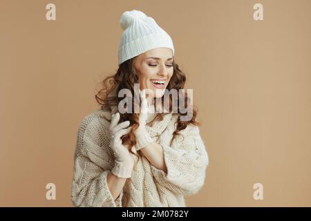 Ciao inverno. donna sorridente elegante di 40 anni in maglione beige, guanti e cappello su sfondo beige che mostra i capelli. Foto Stock