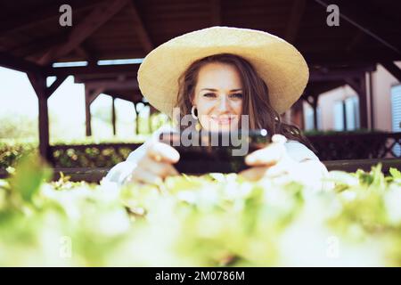 donna moderna sorridente di mezza età in camicia bianca con cappello che invia un messaggio di testo utilizzando lo smartphone sul ranch. Foto Stock