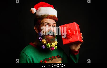 Uomo felice in cappello di Santa e barba decorata con regalo di Natale. Nuovo anno presente box. Foto Stock