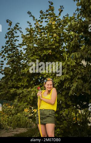 giovane ragazza graziosa che si diverte nel giardino innaffiare le piante con un tubo. Sorridendo mentre prende un hobby favorito Foto Stock