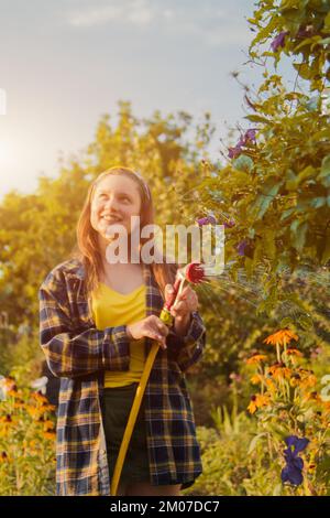 giovane ragazza graziosa che si diverte nel giardino innaffiare le piante con un tubo. Sorridendo mentre prende un hobby favorito Foto Stock