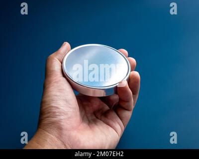 Mano in primo piano con stemma vuoto in acciaio inossidabile di forma rotonda isolato su sfondo blu. Bottone in metallo di forma circolare, color argento e me Foto Stock