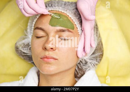 Il concetto di cosmetologia. Primo piano di un estetista che applica una maschera verde con un pennello sul viso di una donna Foto Stock