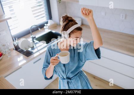 Giovane donna che ascolta musica e che si gode una tazza di caffè al mattino, in cucina. Foto Stock