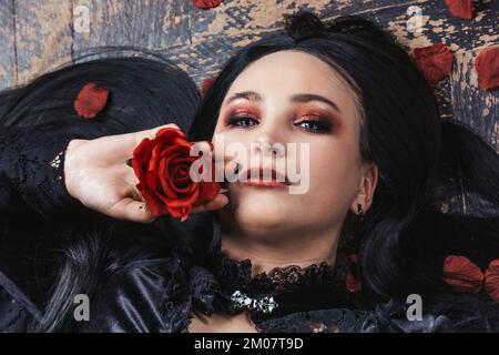 Un modello felice più taglia in un vestito nero con capelli lunghi si trova su un pavimento di legno. Giovane donna in posa su macchina fotografica in petali di rosa. Il corpo è positivo Foto Stock