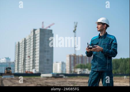 Un uomo in un casco e tute controlla un drone in un cantiere. Il costruttore esegue la supervisione tecnica. Foto Stock