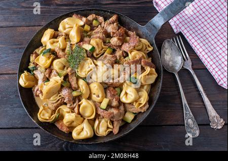 Tortellini con carne, zucchine e salsa di panna in una rustica padella di ferro isolata su tavola di legno. Vista dall'alto Foto Stock