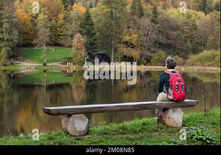 Donna anziana con zaino, migliore Ager, seduta sola su panca di legno, Ebnisee, Kaisersbach, autunno, Foresta sveva, Baden-Württemberg, Germania, Europa Foto Stock