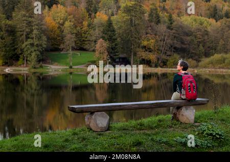 Donna anziana con zaino, migliore Ager, seduta sola su panca di legno, Ebnisee, Kaisersbach, autunno, Foresta sveva, Baden-Württemberg, Germania, Europa Foto Stock