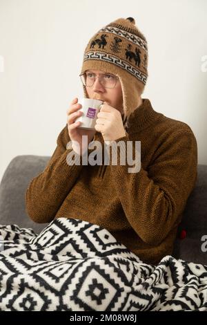 Un giovane uomo che indossa un cappello e un maglione di lana e che beve un tè alle erbe Foto Stock