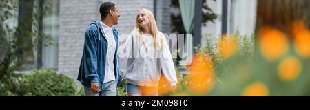 donna bionda felice che parla con il ragazzo afro-americano mentre cammina in città su primo piano sfocato, banner Foto Stock