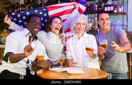 Allegri tifosi multirazziali che sventolano la bandiera degli Stati Uniti mentre bevono birra e guardano il torneo nello sport bar Foto Stock