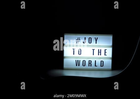 Scatola luminosa con la frase "# Joy to the World" illuminata su di essa posta in ombra e sfondo scuro Foto Stock