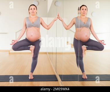 Donna incinta è impegnata nello yoga. Posa dell'albero o Vrikshasana, tenendo con la mano vicino alla parete dello specchio Foto Stock