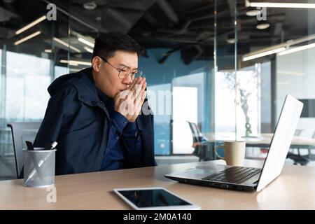 Non c'è riscaldamento in ufficio e il riscaldamento è rotto, l'uomo d'affari si congela sul posto di lavoro, l'uomo è seduto al tavolo all'interno dell'edificio utilizzando un computer portatile al lavoro. Foto Stock