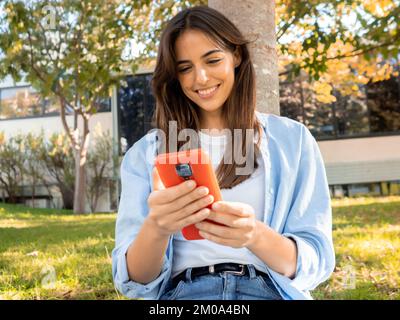 Giovane donna sorridente che chiacchiera con uno smartphone in un parco Foto Stock