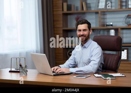 Ritratto di un investitore di successo in ufficio, uomo maturo sorridente e guardando la fotocamera utilizzando un computer portatile al lavoro e indossando cuffie bianche per videochiamare, Foto Stock