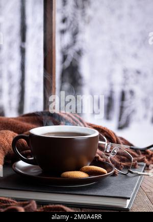 Una tazza calda fumante di caffè o tè con un libro e una coperta accanto a una finestra con sfondo invernale innevato Foto Stock