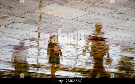 Astratto blurry silhouette ombra riflessi di persone irriconoscibili riflessioni a piedi sul bagnato strada della città strada marciapiede in un giorno di primavera piovoso Foto Stock