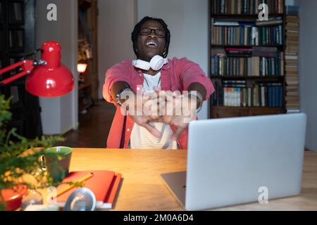 Ragazzo afro addormentato che sbadigia le braccia mentre studia sul computer portatile per lunghe ore a casa Foto Stock
