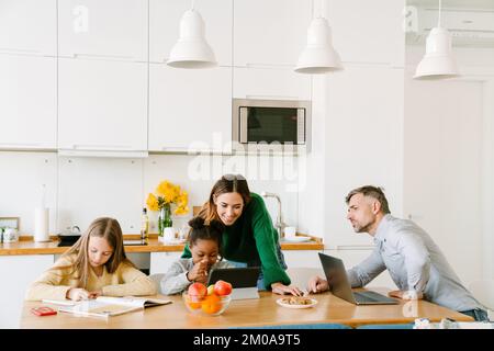 Famiglia felice sulla cucina accogliente a casa, madre e padre lavorante aiutare le loro figlie con i compiti a casa Foto Stock