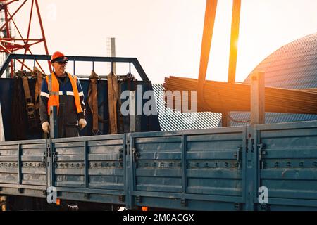 Infilare il casco e il giubbotto scarica tubi metallici e raccordi dalla carrozzeria del camion in una giornata limpida. Background di produzione. Flusso di lavoro autentico in cantiere. Slinger al lavoro.. Foto Stock