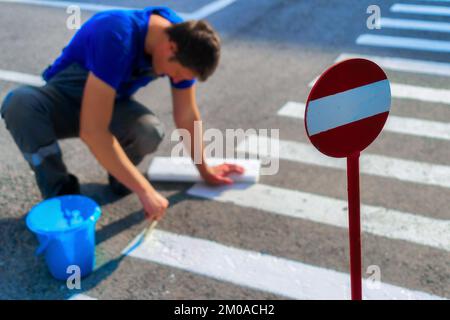 L'operaio del servizio stradale si siede sul suo grembo e dipinge pedone a strisce incrociando su asfalto con pennello. Marcature per pedoni sulla carreggiata. Transizione sicura per le persone. Flusso di lavoro reale... Foto Stock