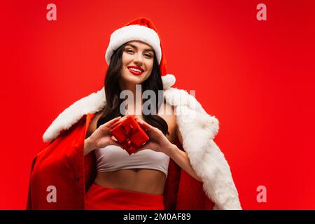 Donna brunette sorridente in cappello di santa e costume che tiene scatola regalo isolato su rosso Foto Stock