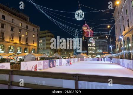 Piazza con pista di pattinaggio su ghiaccio e mercatino di Natale con luci colorate, bancarelle e grande albero di Natale di notte. Foto Stock