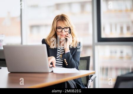 Donna d'affari di mezza età che utilizza un computer portatile ed effettua una chiamata mentre si siede alla scrivania in ufficio. Donna d'affari executive che indossa occhiali e blazer. Foto Stock