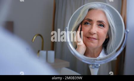 Pensivo 50s donna caucasica di mezza età senior pelle toccare il viso guardando specchio riflessione sorridente godere morbido viso. Signora anziana coccole sano Foto Stock