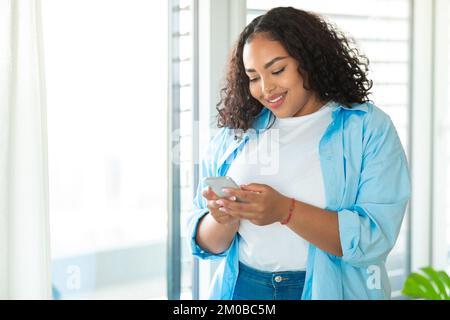 Donna obesa nera felice usando il cellulare e il testo, in piedi vicino alla finestra a casa, copia spazio Foto Stock