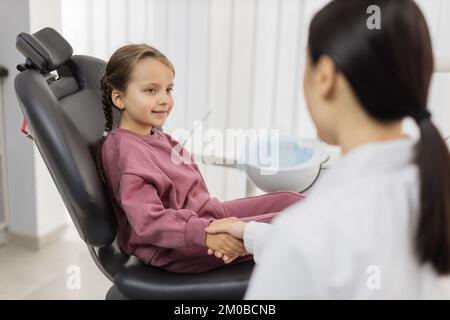 Giovane donna pediatrica dentista scuotendo cute poco sorridente bambina paziente mano dopo il trattamento dei denti, curando con successo in clinica dentale moderna e luminosa. Foto Stock