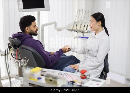 Giovane odontoiatra pediatrica asiatica che scuote la mano del paziente, uomo sorridente, dopo il trattamento dei denti, curandosi con successo in clinica odontoiatrica moderna e luminosa. Foto Stock