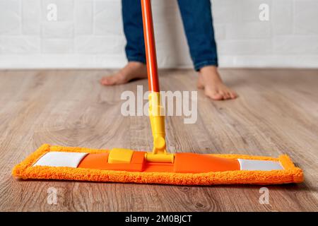 Donna a piedi nudi che muta il pavimento. Pavimento in laminato di legno pulente femmina con mocio in microfibra arancione a casa. Routine di lavoro, pulizia, pulizia. Foto Stock