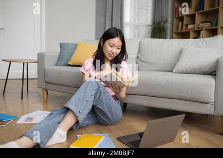 Felice giovane signora giapponese che studia online da casa, leggendo un libro e usando un pc portatile, seduto sul pavimento nel soggiorno Foto Stock