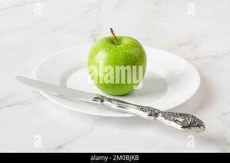 Mela verde fresca e coltello su un piatto su sfondo di marmo. Frutta matura e succosa per un'alimentazione sana e antiossidante. Spuntino vegetariano vitaminico. Vista frontale. Foto Stock