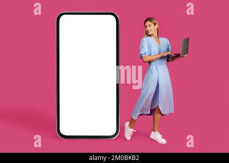 Sorridente bella donna adulta caucasica bionda in abito con notebook guarda enorme smartphone con schermo vuoto Foto Stock