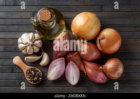 Salotti rossi, cipolle gialle, aglio, pepe e olio d'oliva su fondo di legno nero. Bulbi di cipolla freschi interi e tagliati a metà. Spezie e condimenti Foto Stock