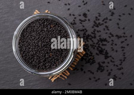 Vaso di vetro pieno di semi di cumino nero su sfondo nero. Macro. Cucina con semi nigella concetto. Kalonji come spezie e condimento. Nigella sativa. Foto Stock