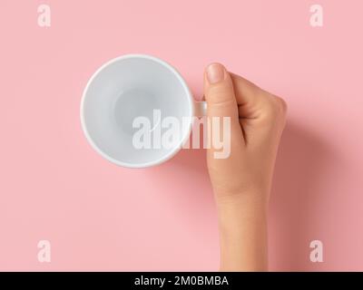La mano della donna tiene la tazza vuota sopra lo sfondo rosa. Femmina caucasica che tiene una tazza da tè bianca. Bevande calde ed elemento di design per la colazione. Vista dall'alto. Foto Stock