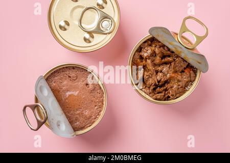 Cibo gatto bagnato in vasi aperti su sfondo rosa pastello. Mangime per animali domestici umido in lattine di metallo primo piano. Pezzi di carne in scatola e pate molle per animali domestici. Foto Stock