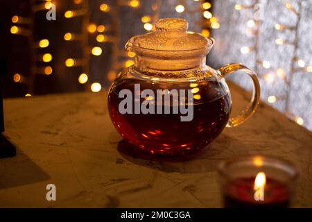 bollitore in vetro fotografico con tè e candela su sfondo grigio irregolare sullo sfondo di un albero di Natale in fiamme Foto Stock