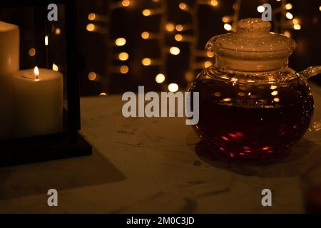 teiera in vetro fotografico con tè accanto a una candela e luci accese sullo sfondo Foto Stock