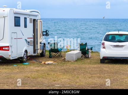 Camper parcheggiata con il mare sullo sfondo e due sedie con vista mare. Turista che riposa sulla sedia accanto al suo cane su un guinzaglio. Un gabbiano Foto Stock