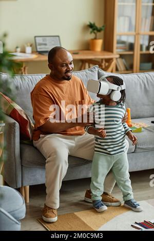 Ritratto verticale di un simpatico bambino nero che indossa un visore VR e ti offre l'intrattenimento in realtà virtuale con il supporto del padre Foto Stock