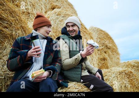 Due giovani lavoratori felici di fattoria in abbigliamento da lavoro seduto da pila di fieno di fronte alla macchina fotografica, parlando e avendo panini e tè per pranzo Foto Stock