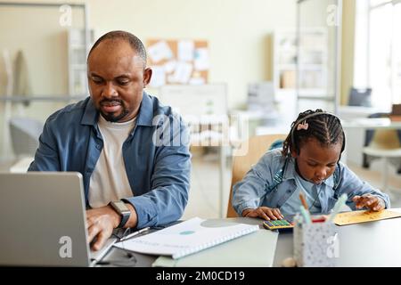 Vista frontale ritratto di un singolo padre nero che lavora da casa con un bambino che gioca con la calcolatrice accanto a lui Foto Stock