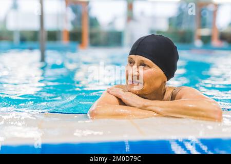 Pensiva nonna caucasica in berretto nero capelli rilassarsi sul lato della piscina interna. Pensionati attivi. Foto di alta qualità Foto Stock